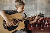A young boy sitting indoors playing an acoustic guitar on a sofa.